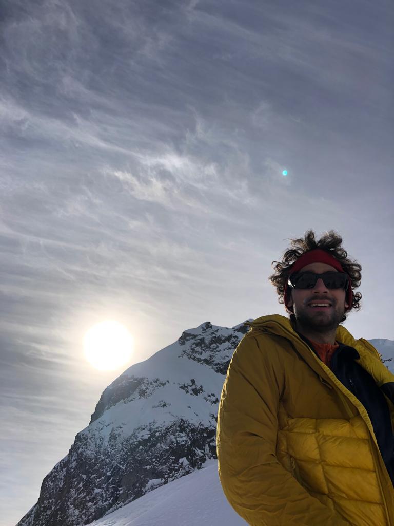 Hadrien Saiag chercheur en anthropologie économique souriant, en tenue d'alpiniste, devant une montagne au coucher du soleil